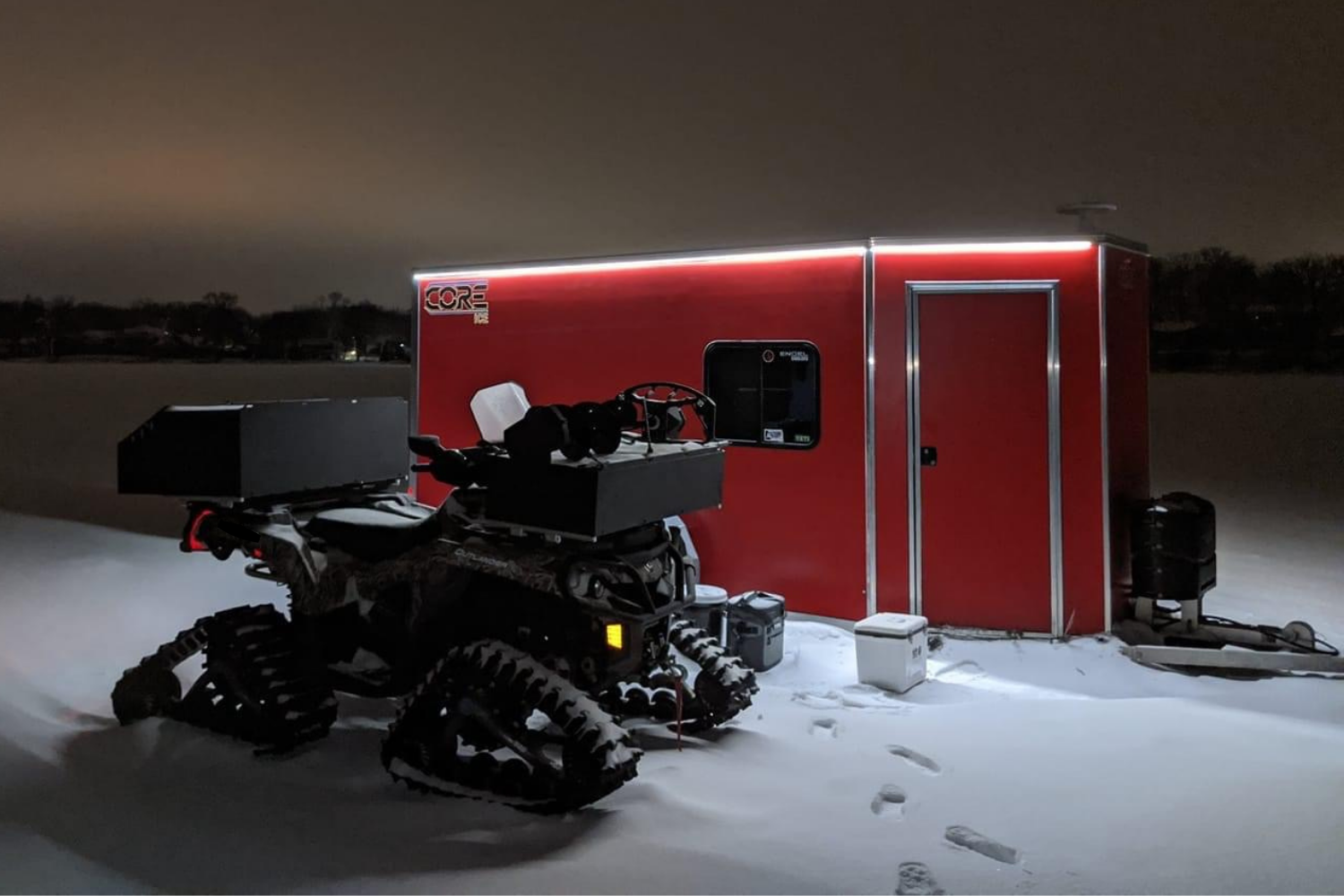Red CORE ICE trailer set outside on the ice at night behind a four wheeler with tracks on it, a cooler set out front and the LED lights on
