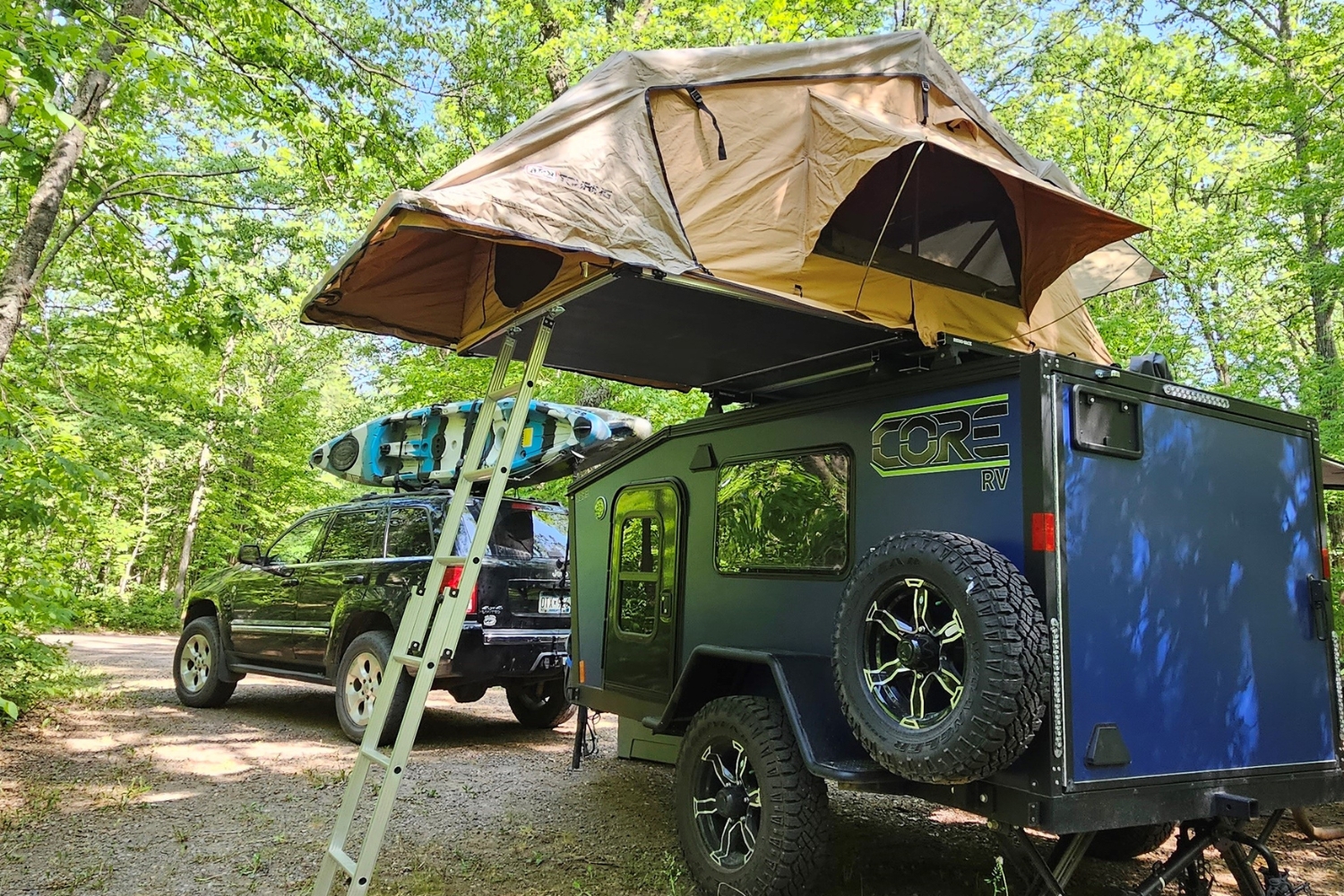 Exterior shot of a blue CORE RV 5510 pulled behind a jeep with mounted kayaks backed into a campsite with a rooftop tent open and ladder down.
