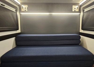 Interior view of the CORE RV 5510 rear interior with foldable sofa (queen bed) and upper storage with side cubbies and a passthrough to the back galley