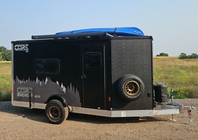Exterior view of the CORE RV 6515 Trailhead curbside with grey tree graphics and awning on the roof and a mounted kayak